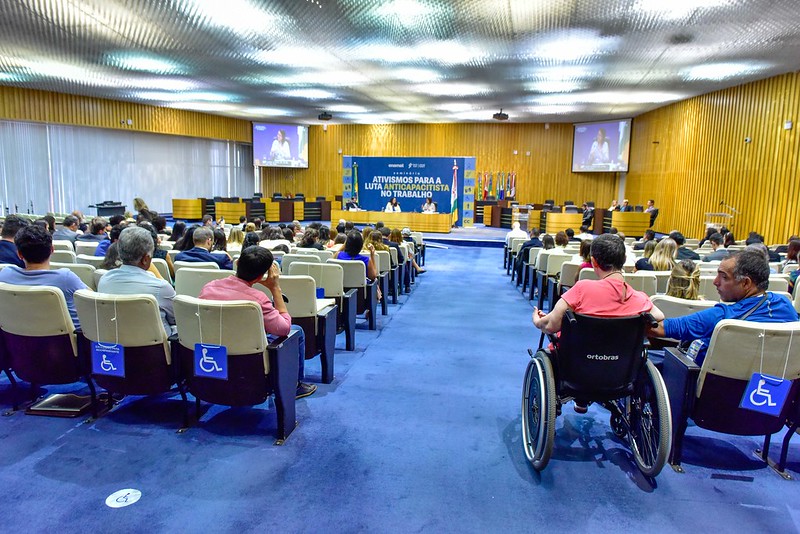 Foto do plenário onde foi realizado o evento. Ao fundo, a mesa de apresentações e em primeira perspectiva pessoas assistindo, incluindo uma pessoa cadeirante. O local tem carpete azul, paredes em ripas de madeira amarelada e teto luminoso prateado.