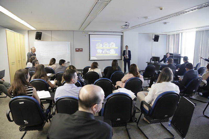 Foto do sala onde foi promovido o encontro. Há uma pessoa em pé ao lado do datashow e pessoas sentadas em cadeiras.