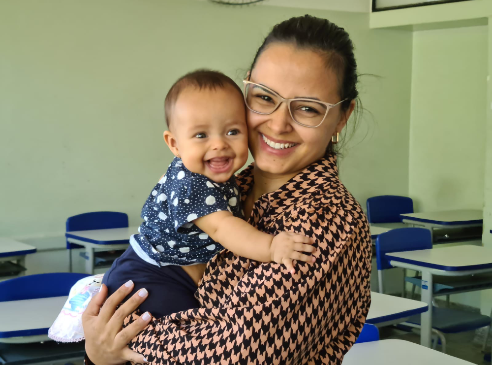 Advogada Maíra Maria Soares Shirasu segurando a filha no colo. A mulher usa óculos, cabelo amarrado e blusa xadrez. A bebê sorri e está com uma blusa azul. 