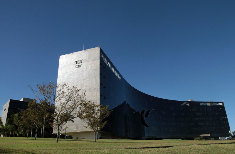 Foto do edifício-sede do TST e do CSJT em perspectiva diagonal de baixo pra cima em um céu azul e sem nuvens.