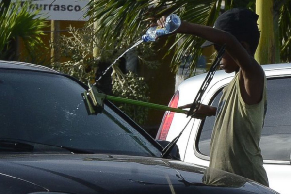 Menino negro jogando agua em para-brisas de um carro. Ele usa uma garrafa de plástico e rodo.