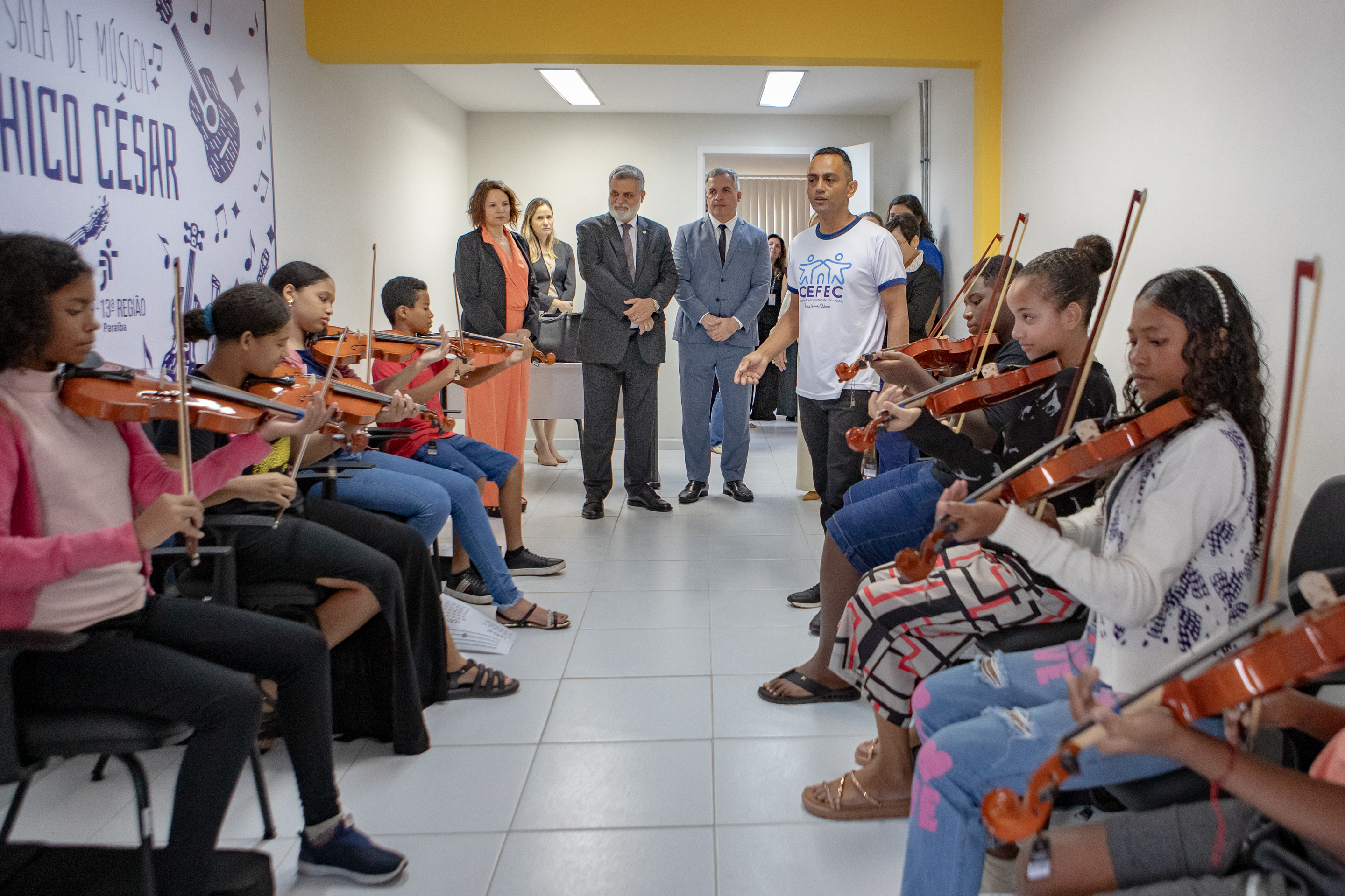 Ministro e o presidente do TRT-13 observação uma apresentação de jovens com violinos.